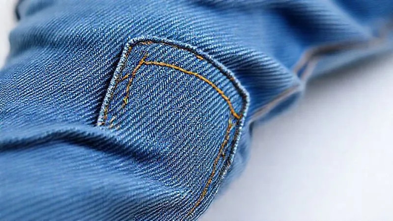A photorealistic close-up, macro shot of a pair of children's denim jeans, focusing on a reinforced knee patch with visible double-stitching and a strong flat-felled side seam. The fabric texture is clear, showing subtle wear but robust construction. Professional photography, 8K, cinematic lighting, extremely sharp focus on the stitching and seam, with a very shallow depth of field.