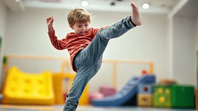 A photorealistic shot from a slightly elevated angle of a child, approximately 7 years old, enthusiastically testing the flexibility and durability of a pair of pants by doing a playful high-kick in a brightly lit, modern indoor play area. The child's clothing appears well-made and allows full range of motion. A blurred background shows soft play equipment. Professional photography, 8K, cinematic lighting, sharp focus on the child, capturing dynamic movement.
