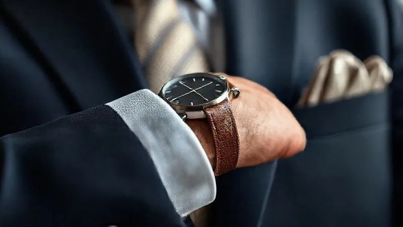 A photorealistic, professional photography close-up of an executive's wrist, featuring a modern minimalist watch with a unique leather strap, peeking out from a crisp suit cuff. In the background, a subtly textured silk tie and a folded pocket square are visible. Cinematic lighting, sharp focus on the watch and cuff, depth of field blurring the background, 8K hyper-detailed, shot on a high-end DSLR.
