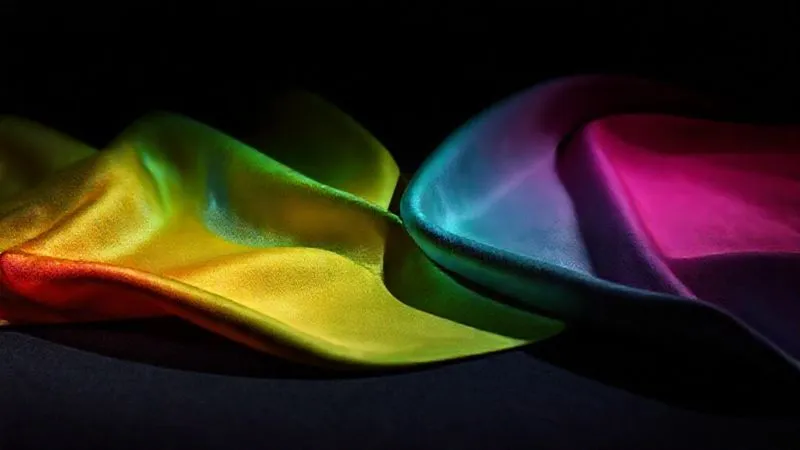 A photorealistic shot of various fabric swatches (polyester satin, mercerized cotton, theatrical velvet, silk) arranged on a dark surface, with dramatic stage lighting illuminating them from different angles. One swatch shows vibrant color, while another shows subtle fading under the same light. Professional photography, 8K, cinematic lighting, sharp focus, depth of field, shot on a high-end DSLR.