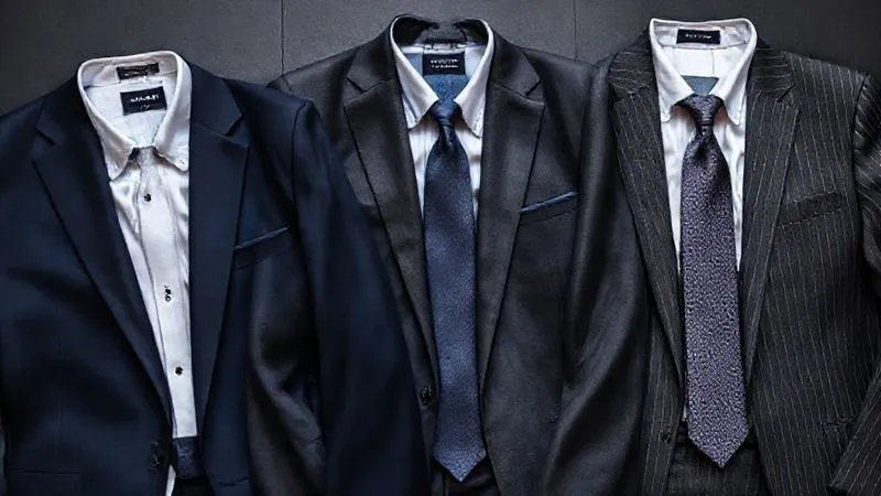 A photorealistic, professional photography, 8K, cinematic lighting, sharp focus, depth of field, shot on a high-end DSLR, showcasing a flat lay of three impeccably tailored men's suits: one in deep navy, one in charcoal grey, and one with a subtle pin-stripe pattern. Each suit includes a crisp white shirt and a complementary silk tie, emphasizing strategic color and pattern choices for professional authority.