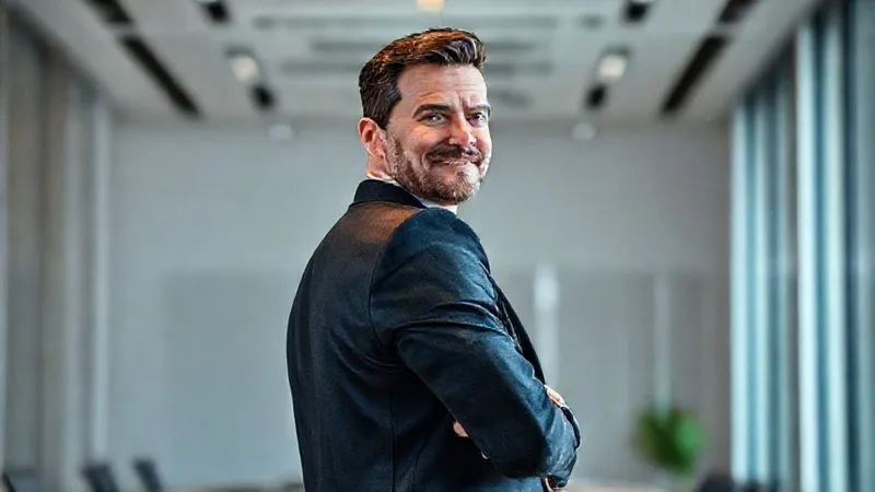 A photorealistic, professional photography, 8K, cinematic lighting, sharp focus, depth of field, shot on a high-end DSLR, of a confident businessman in a tailored suit, standing tall with shoulders back, making direct eye contact, and a subtle, reassuring smile. The background is a modern, minimalist office, emphasizing his strong, authoritative posture.