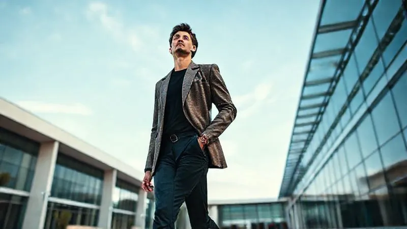 A photorealistic, professional photography shot of a stylish non-model captured from a slightly low angle, walking confidently and looking upwards towards the sky with a thoughtful expression. The background features modern architecture and a clear blue sky. Cinematic lighting, sharp focus on the subject, 8K hyper-detailed.