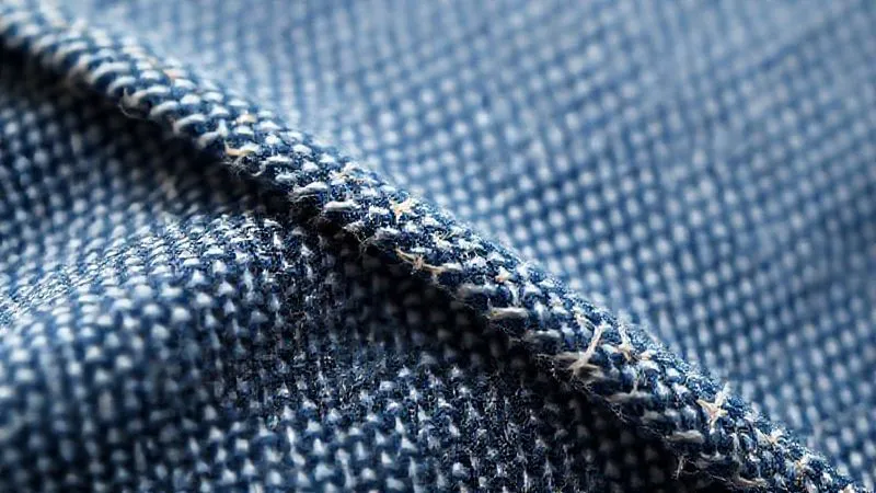 A close-up photorealistic image of a woven natural fiber fabric, possibly a denim-like twill, with subtle, strategically placed reinforcement zones visible as slightly denser or differently patterned sections. The texture is rich and detailed, showing individual threads. The lighting is strong, highlighting the structural integrity. Professional photography, 8K, cinematic lighting, sharp focus, depth of field, shot on a high-end DSLR.