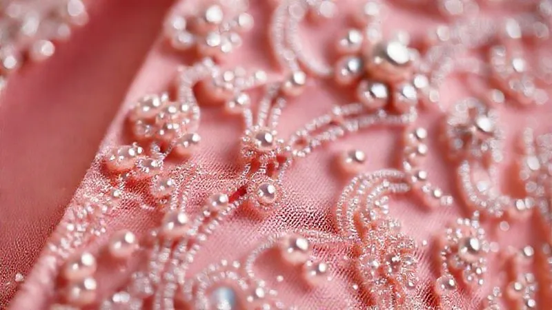 A photorealistic, professional photography, 8K close-up of intricate hand-stitching and delicate beadwork on a haute couture garment, highlighting the exquisite detail, texture, and shimmering quality of the embellishments. Cinematic lighting, sharp focus, extreme depth of field to emphasize the craftsmanship, shot on a high-end DSLR.