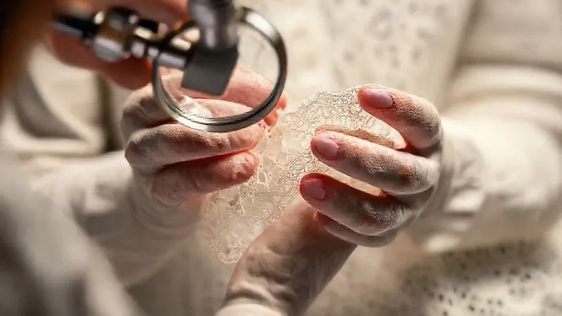 Expert hands gently examining a delicate, heavily damaged vintage lace textile under a magnifying glass, in a well-lit textile conservation studio, photorealistic, professional photography, 8K, cinematic lighting, sharp focus on the hands and fabric, depth of field, shot on a high-end DSLR.