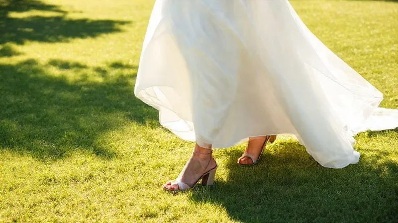 A photorealistic image of a bride confidently walking on a soft, sun-drenched lawn, her elegant, practical block heel bridal shoes making stable contact with the grass. Her gown flows gracefully, and she looks ahead with a serene smile. Professional photography, 8K, cinematic lighting, sharp focus, depth of field, shot on a high-end DSLR, conveying grace and ease.