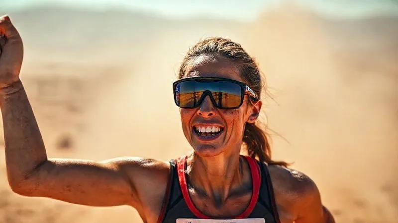A triumphant, photorealistic image of a female ultra-marathoner crossing a finish line in a desert landscape, her face streaked with sweat but her high-performance sports eyewear perfectly in place. Her expression is one of exhaustion and elation, showcasing the secure fit of her gear under extreme conditions. Dust and heat haze in the background, cinematic lighting, 8K, sharp focus, depth of field, professional photography, shot on a high-end DSLR.