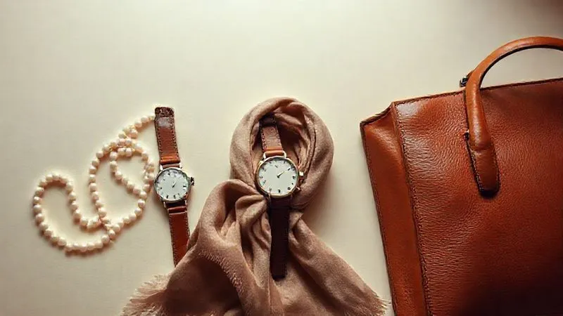 A curated display of iconic classic accessories including a string of pearls, a vintage leather watch, a folded silk scarf, and a structured leather handbag, arranged aesthetically on a smooth, neutral surface under soft, diffused lighting. Photorealistic, professional photography, 8K, cinematic lighting, sharp focus, depth of field.