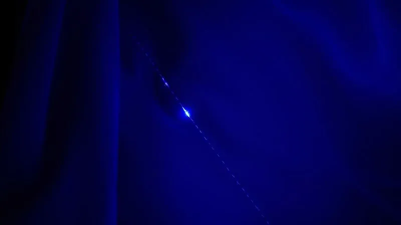 Photorealistic, professional photography, 8K, cinematic lighting, sharp focus, depth of field, shot on a high-end DSLR. A close-up of a delicate 1920s silk flapper dress fabric under a UV blacklight. The silk glows with a subtle, consistent luminescence, but a small, hidden repair or patch made with modern thread fluoresces distinctly, appearing as a brighter or different colored line, highlighting the contrast. The scene is dark, with the UV light casting an eerie glow on the fabric.
