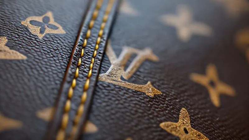 A macro shot, photorealistic, professional photography, of the iconic Louis Vuitton monogram canvas, focusing on the precise alignment of the LV logos across a seam, with a hint of natural vachetta leather developing a honey patina. 8K, cinematic lighting, sharp focus, depth of field.