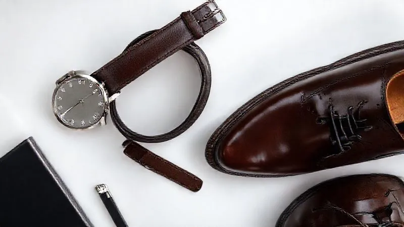 A photorealistic overhead shot of a perfectly coordinated set of men's accessories: a classic silver-toned dress watch with a dark brown leather strap, a dark brown polished leather belt with a subtle silver buckle, and a pair of impeccably shined dark brown leather oxfords. The items are arranged artfully on a minimalist desk, emphasizing their seamless harmony. Professional photography, 8K, cinematic lighting, sharp focus, depth of field, shot on a high-end DSLR.
