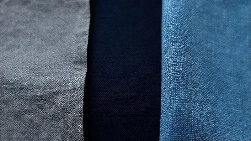A close-up, photorealistic image of three fabric swatches side-by-side: one in solid mid-grey showing a faint damp patch, one in dark navy blue showing no visible dampness, and one in a heathered light blue with a subtle micro-pattern, also showing no visible dampness. Cinematic lighting, sharp focus on fabric texture, depth of field, 8K, shot on a high-end DSLR.