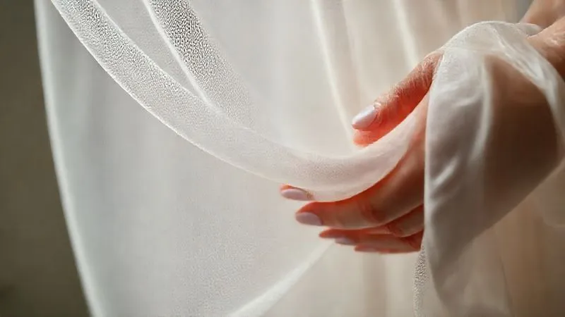A close-up, photorealistic image showcasing the intricate weave of a soft, flowing chiffon fabric, with a subtle texture that hints at its wrinkle-resistant properties. Cinematic lighting highlights individual threads, sharp focus on the fabric, depth of field blurring a bride's hand gently touching it. Shot on a high-end DSLR, 8K hyper-detailed.