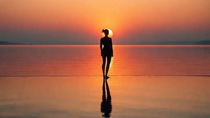 A photorealistic image of a sunrise over a calm, reflective body of water, with a single, elegant fashion silhouette standing on the shore, symbolizing new beginnings, hope, and resilience, cinematic lighting, sharp focus on the silhouette and reflection, depth of field blurring the distant horizon, 8K hyper-detailed, professional photography, shot on a high-end DSLR, serene and inspiring mood.