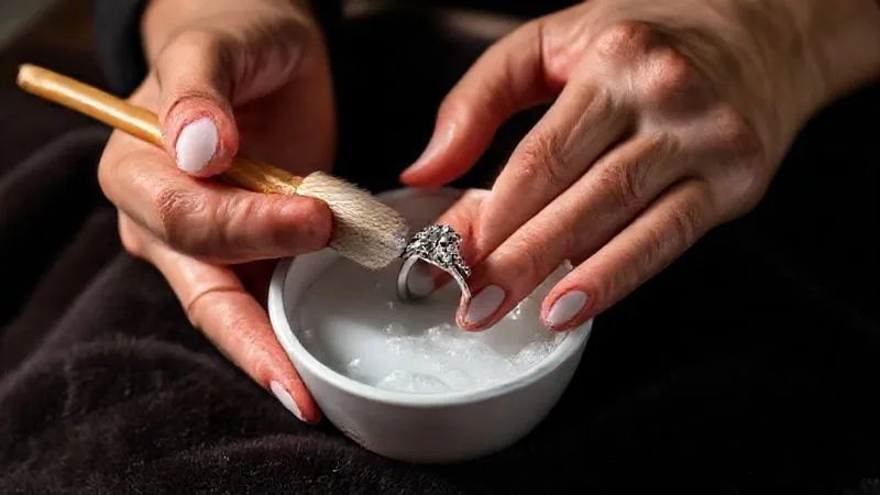 A pair of gentle hands carefully cleaning a sparkling platinum diamond ring with a soft brush and a small bowl of soapy water. The ring sits on a plush, dark velvet cloth. The scene evokes care and maintenance. Photorealistic, professional photography, 8K, cinematic lighting, sharp focus on the hands and ring, depth of field blurring the background, shot on a high-end DSLR.
